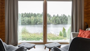 A room with a large window offering a view of a lake and forest. There are two chairs and a small table. - Lakefront Holiday Villa - Traditional Finnish sauna & outdoor jacuzzi (Uurainen)