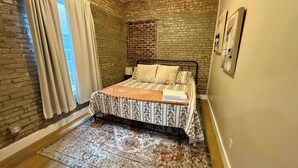A bedroom with a brick wall, a bed with patterned bedding, a window with curtains, and a rug.