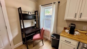 A compact kitchenette with a wooden desk, a chair, a trash bin, a coffee maker, and a window with curtains.