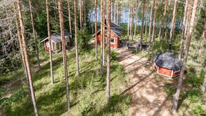 A forested area with a small cabin and a larger building, surrounded by tall trees and a dirt path.