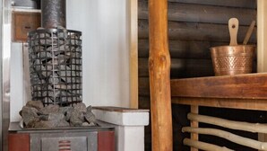 A wood-burning stove with a visible fire, a metal basket on top, and a wooden shelf with a copper pot.