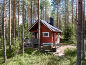 A cabin in a forest with a wooden deck, a hot tub, and a chimney.