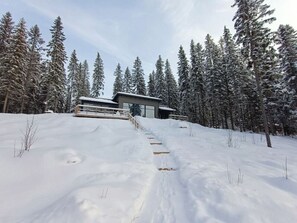 Une maison moderne, avec un paysage enneigé, des marches en bois menant à l’entrée et de grands arbres à feuilles persistantes en arrière-plan.