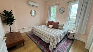 A bedroom with a bed, a bedside table, a small round table, a potted plant, and a window with curtains.
