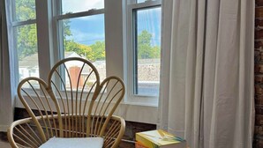 A room with a wicker chair, a small table with books, and a view through large windows.