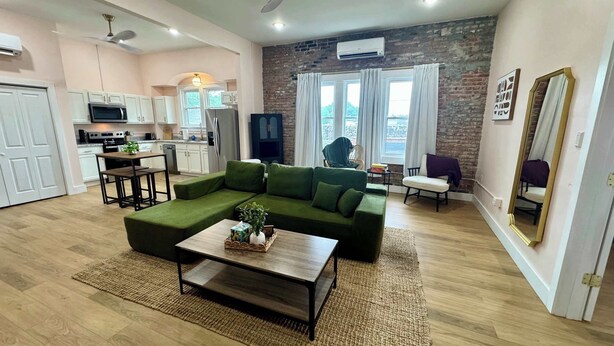 A modern living room with a sectional sofa, a wooden coffee table, and a kitchen area with white cabinets and appliances.