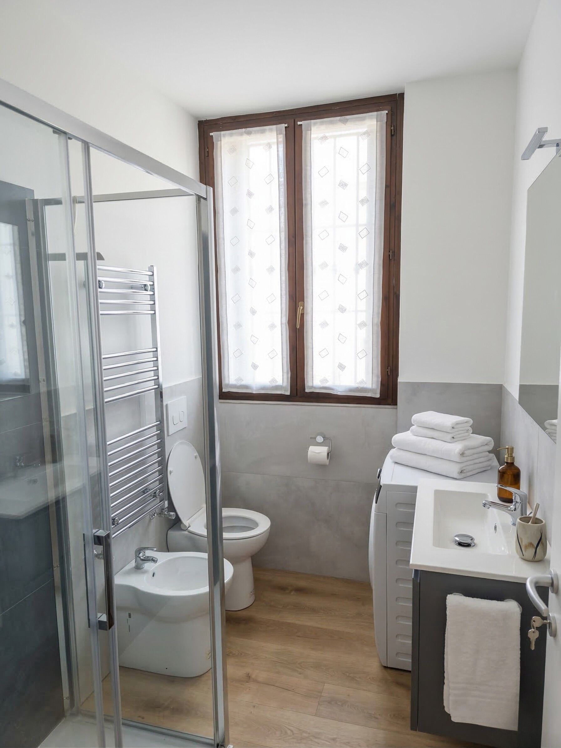 Ein Badezimmer mit Duschkabine aus Glas, Toilette, Waschbecken mit Handtuchhalter und Fenster mit gemusterten Vorhängen.