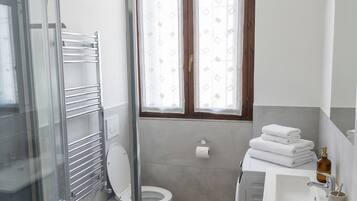 Ein Badezimmer mit Duschkabine aus Glas, Toilette, Waschbecken mit Handtuchhalter und Fenster mit gemusterten Vorhängen.