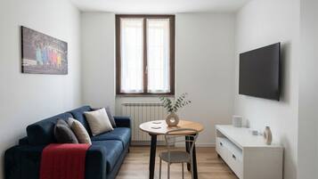 Ein modernes Wohnzimmer mit einem blauen Sofa, einem kleinen runden Tisch, einer weißen Kommode und einem an der Wand montierten Fernseher.