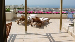 Un balcon avec des fauteuils de jardin en bois, une table et une vue sur la mer et le paysage urbain.