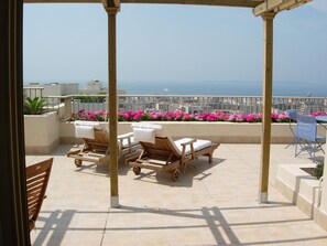 Ein Balkon mit Holzliegen, einem Tisch und Blick auf das Meer und die Stadt.