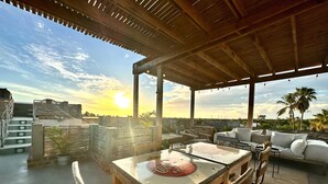 Un espace extérieur aménagé, abrité et doté de tables et de chaises, d’un canapé, et offrant une vue sur le coucher de soleil derrière les palmiers.