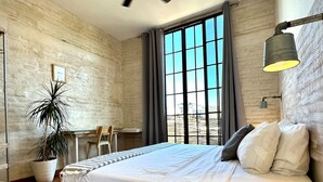 Een slaapkamer met een bed, een bureau, een stoel, een potplant en een raam met gordijnen.