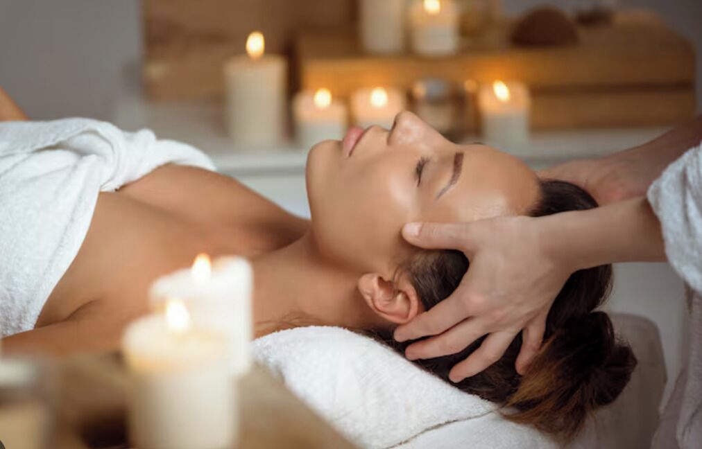 A person receiving a head massage in a spa setting with candles and a wooden shelf in the background.