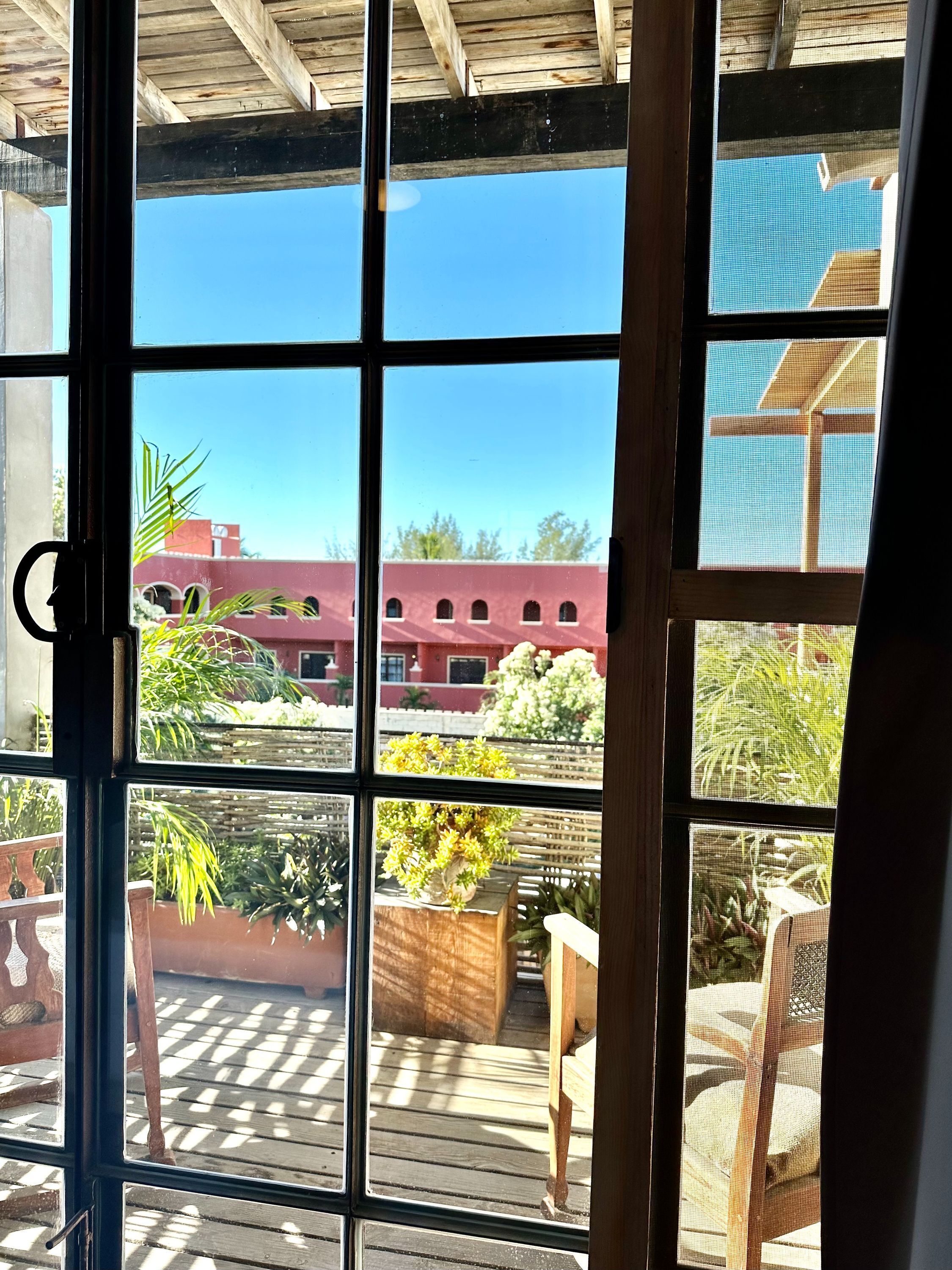 A view through a window with a wooden frame, a red building in the background, and potted plants on the deck.