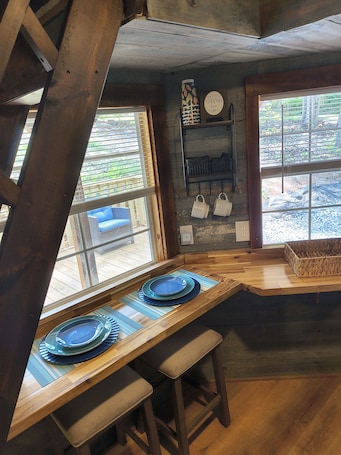 A rustic kitchen with wooden beams, a wooden table set with blue plates, and a window with a view of greenery.. The Moose Lodge, located on Lake Hartwell, in Townville SC. Sleeps 6 guest.