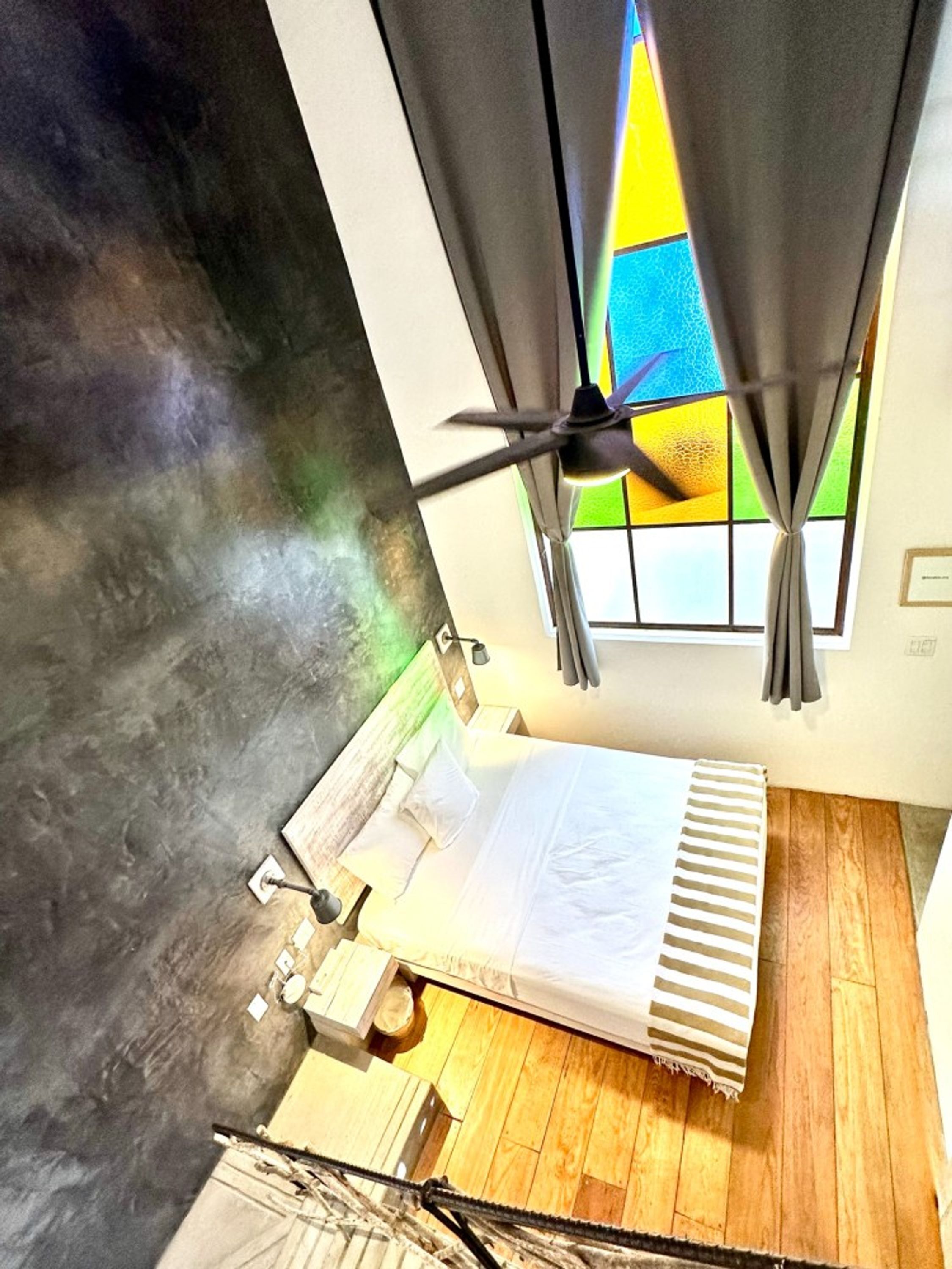 Ein Zimmer mit Bett, einem Vorhangfenster, einem Deckenventilator und Blick auf einen farbenfrohen Außenbereich.