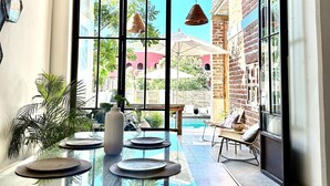 A modern dining area with a glass table, chairs, and a view of a brick building and pool. - TS5 King in Todos Santos (Todos Santos)
