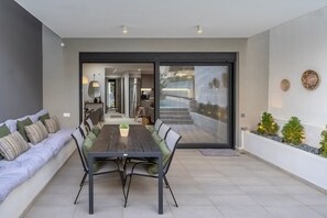 A modern dining area with a wooden table and chairs, a sofa with cushions, and a sliding glass door leading to a kitchen and living space. - Spei Luxury House With Private Pool (Heraklion)