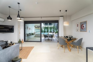 A modern living room with a sofa, dining table, and chairs, a TV, and a view of an outdoor area with a pool. - Spei Luxury House With Private Pool (Heraklion)
