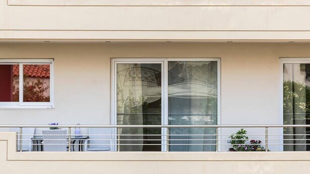 Rakennuksen julkisivussa on suuret ikkunat, parvekkeessa kaide ja parvekkeella on pieni pöytä sekä tuolit.