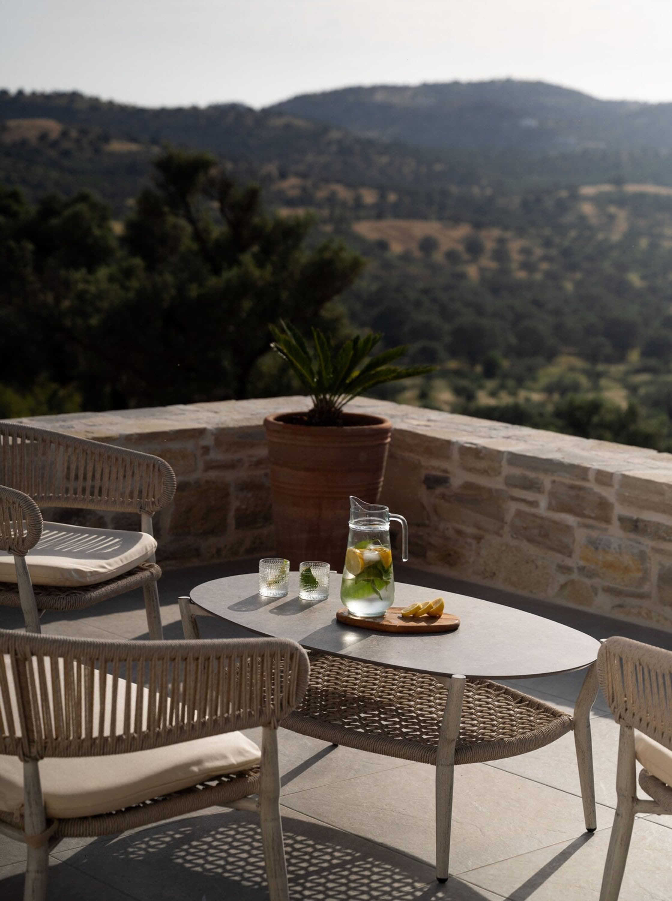 Eine Terrasse mit Korbstühlen, einem runden Tisch, einer Karaffe Wasser mit Zitronenscheiben und einem Blumentopf.