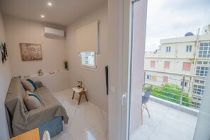 A modern living room with a sofa, a small table, and a chair, a balcony with a view of buildings, and an air conditioning unit on the wall. - Aretousa Apart Heraklion City (Heraklion)