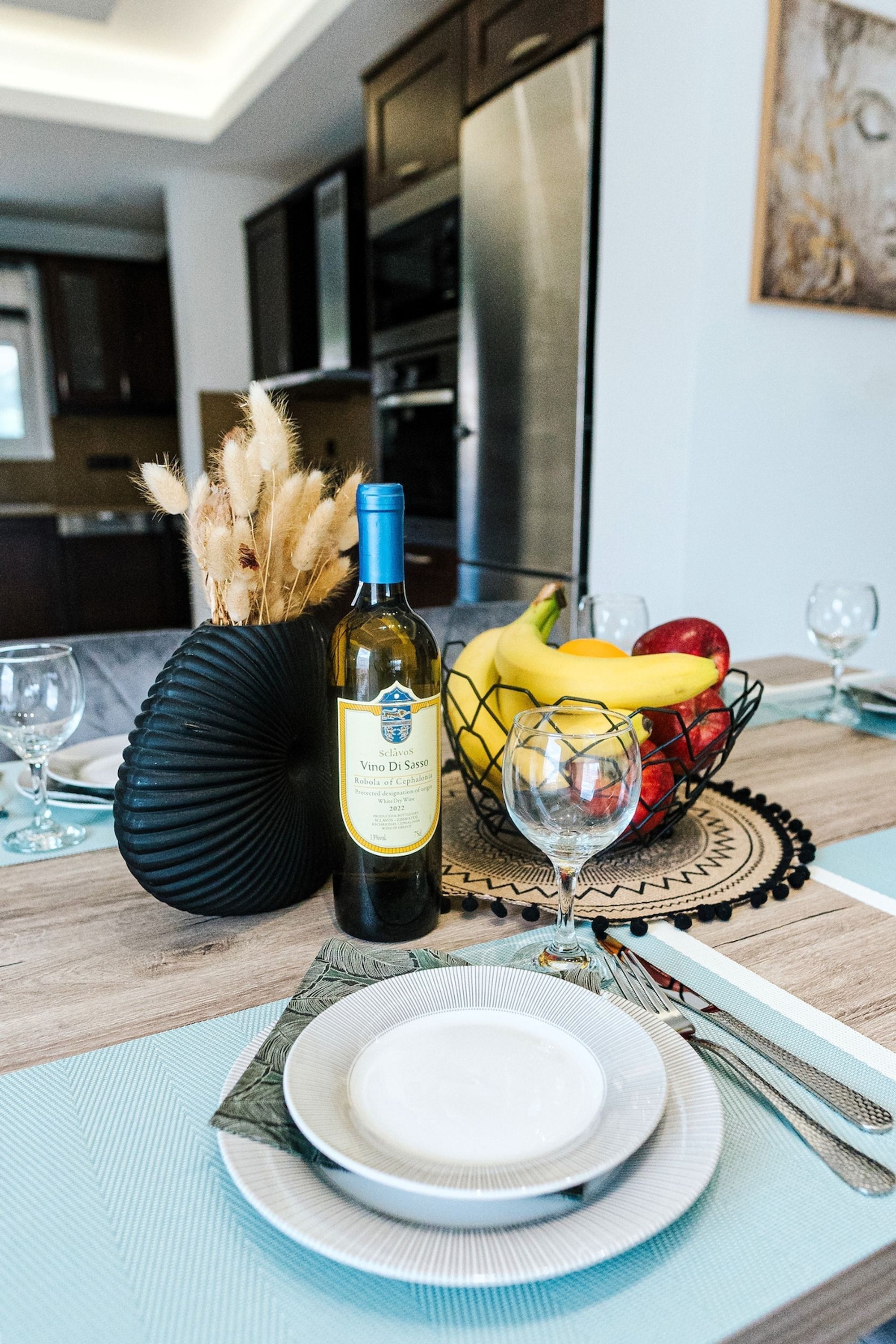 A dining table with a bottle of wine, a fruit bowl, and a vase with dried flowers.