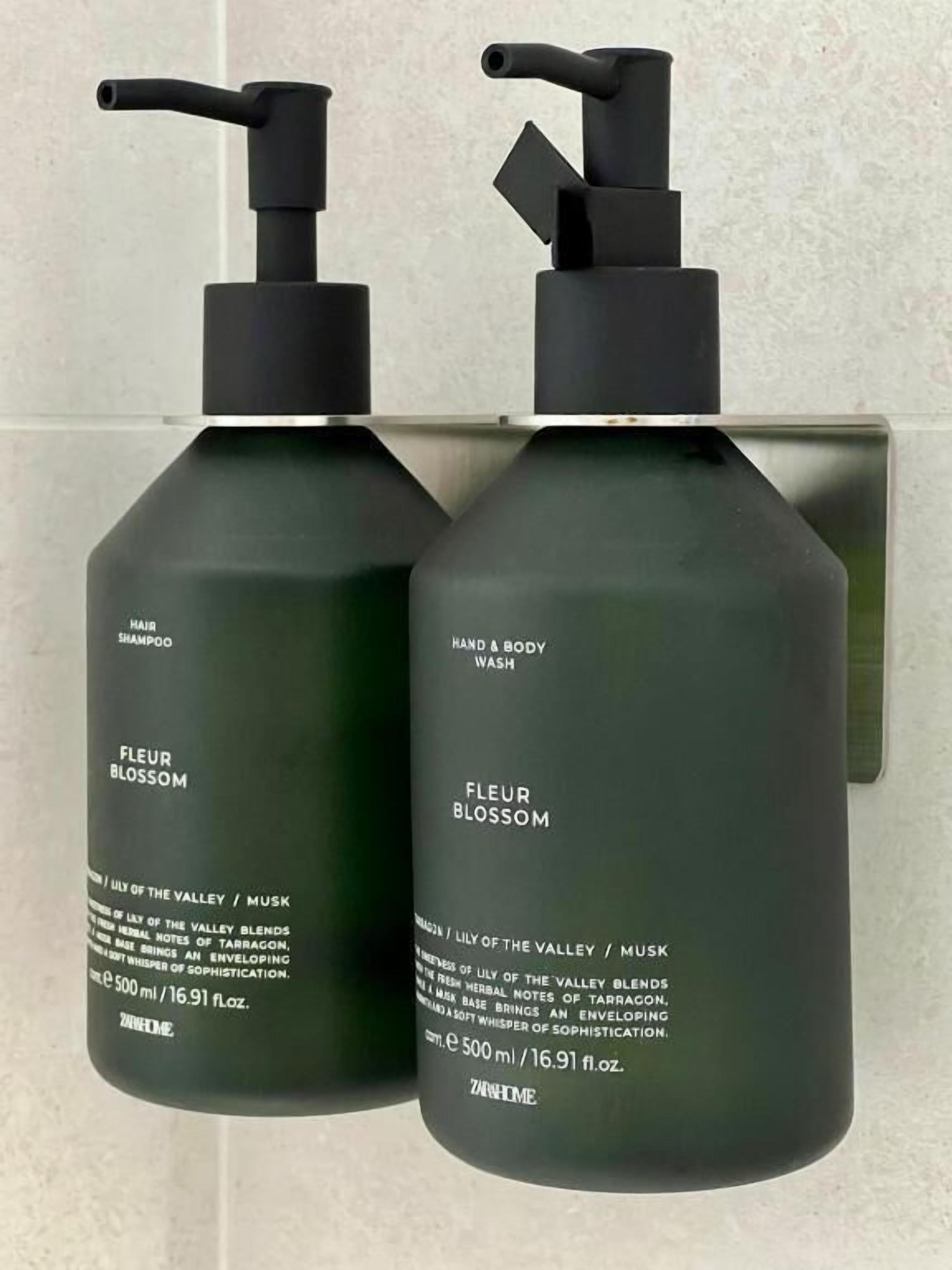 Twee flessen 'Fleur Blossom' shampoo en hand- en bodywash, elk met een pompdispenser, geplaatst tegen een betegelde muur.