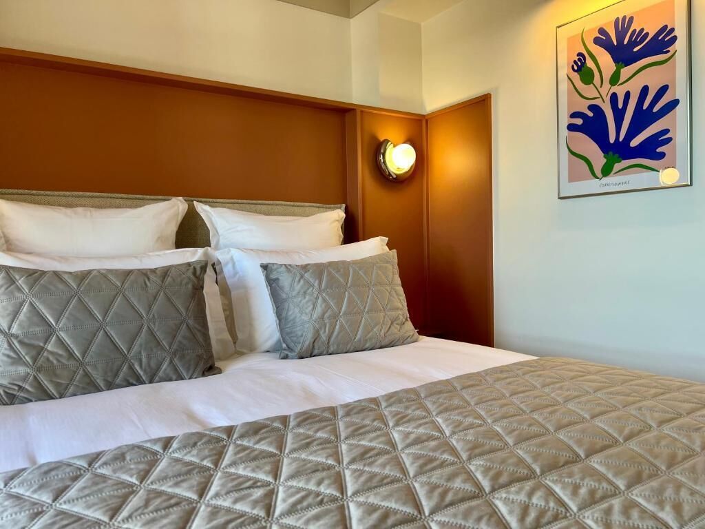 Habitación de hotel con cama, almohadas, luz empotrada en la pared y una obra de arte enmarcada.
