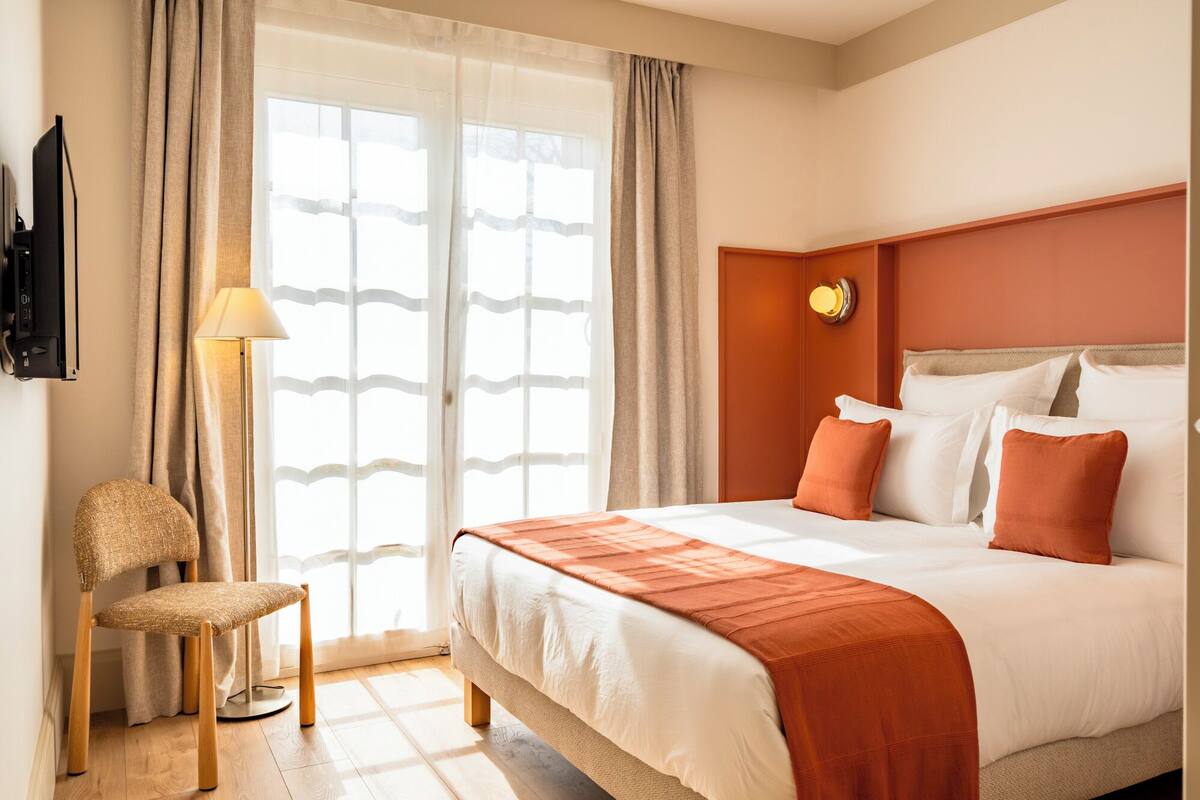 A hotel room with a bed, a chair, a lamp, and a window with curtains.