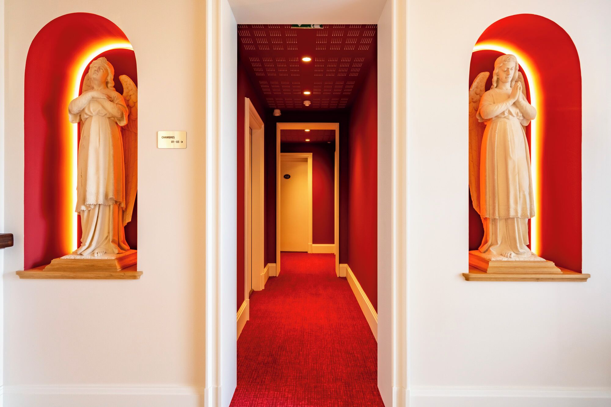Un couloir recouvert d’une moquette rouge, avec deux statues de chaque côté et une porte au fond.