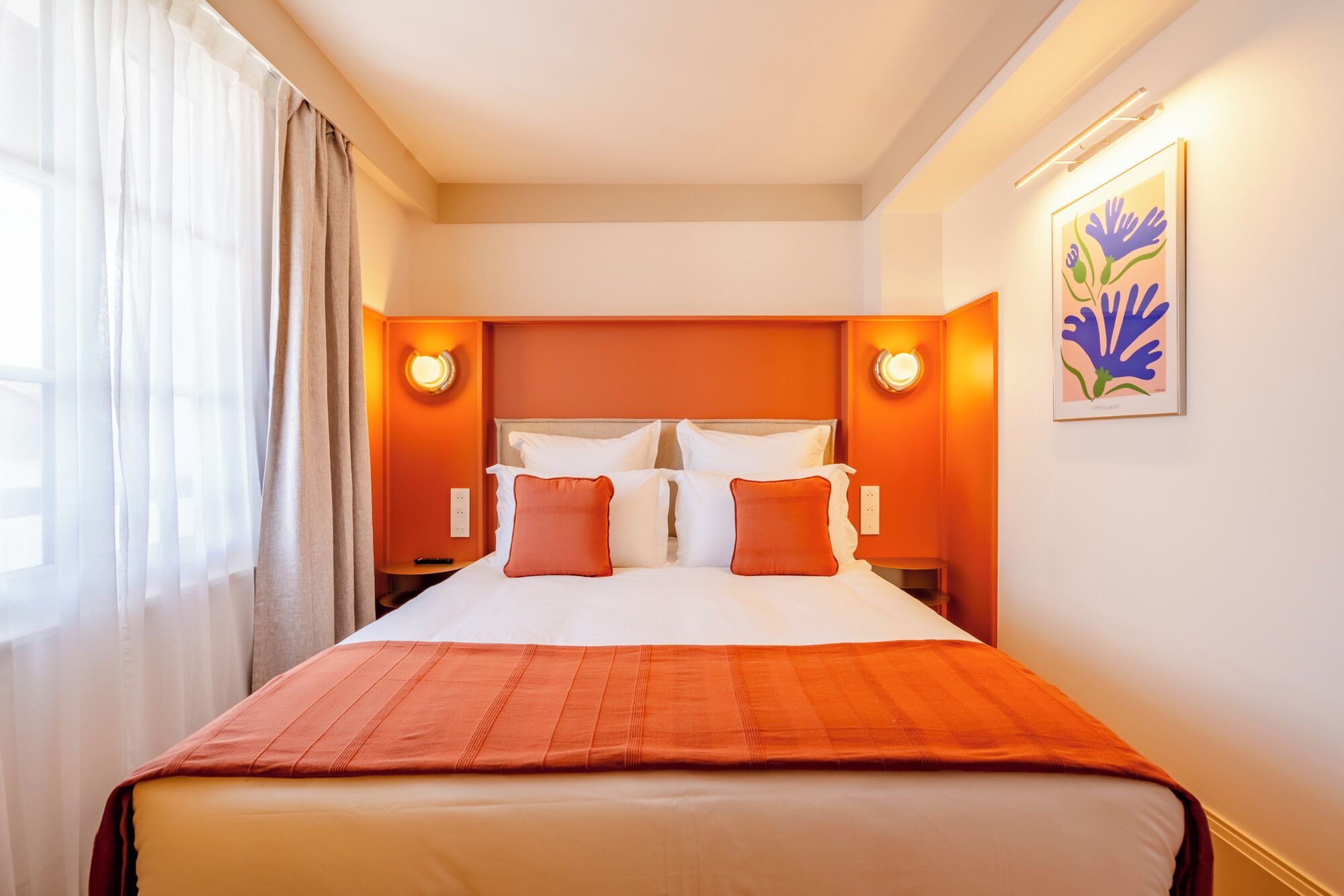 A hotel room with a bed, two bedside tables, a nightstand, a painting on the wall, and a window with curtains.