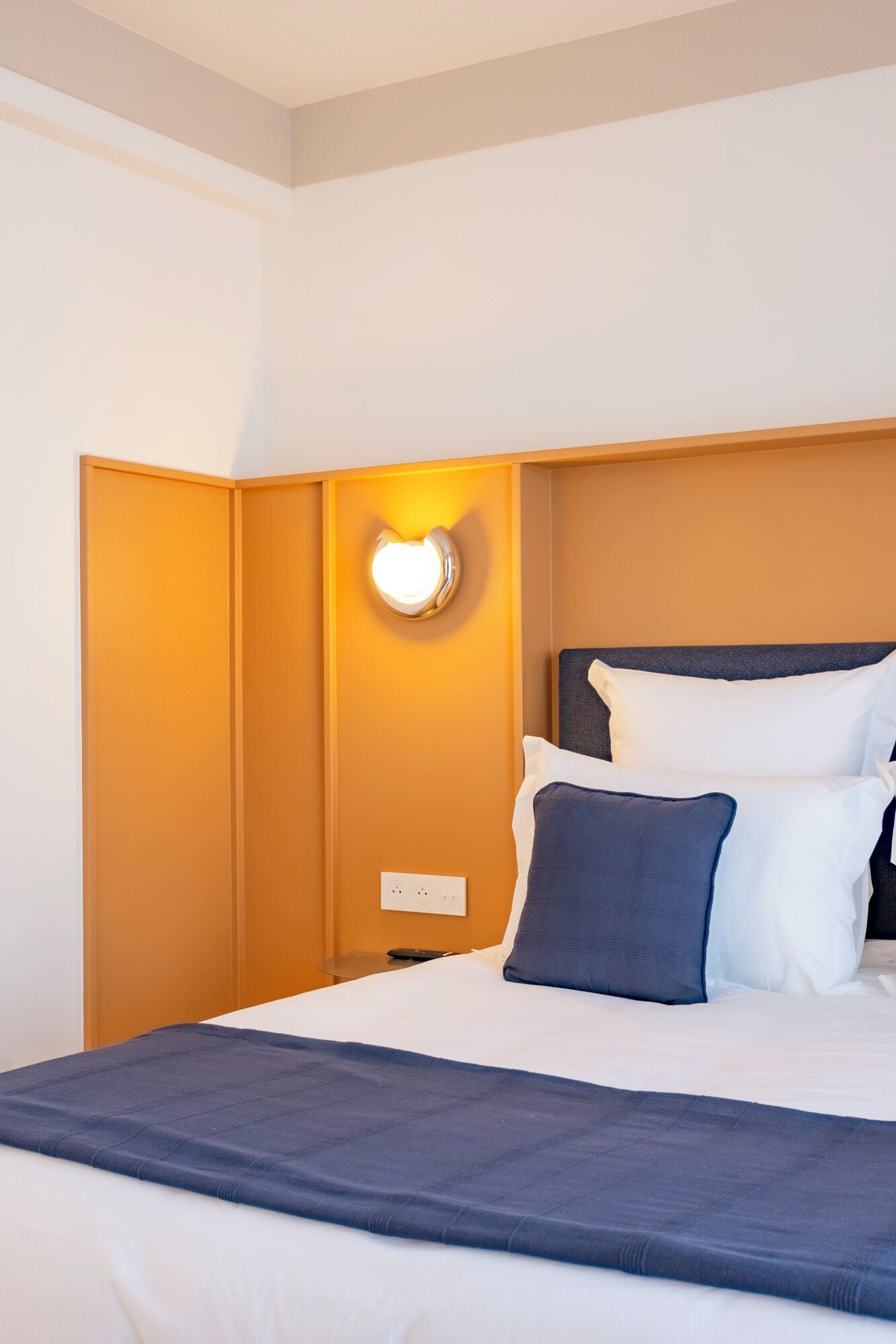 A hotel room with a bed, a nightstand, a wall-mounted light, and a power outlet.
