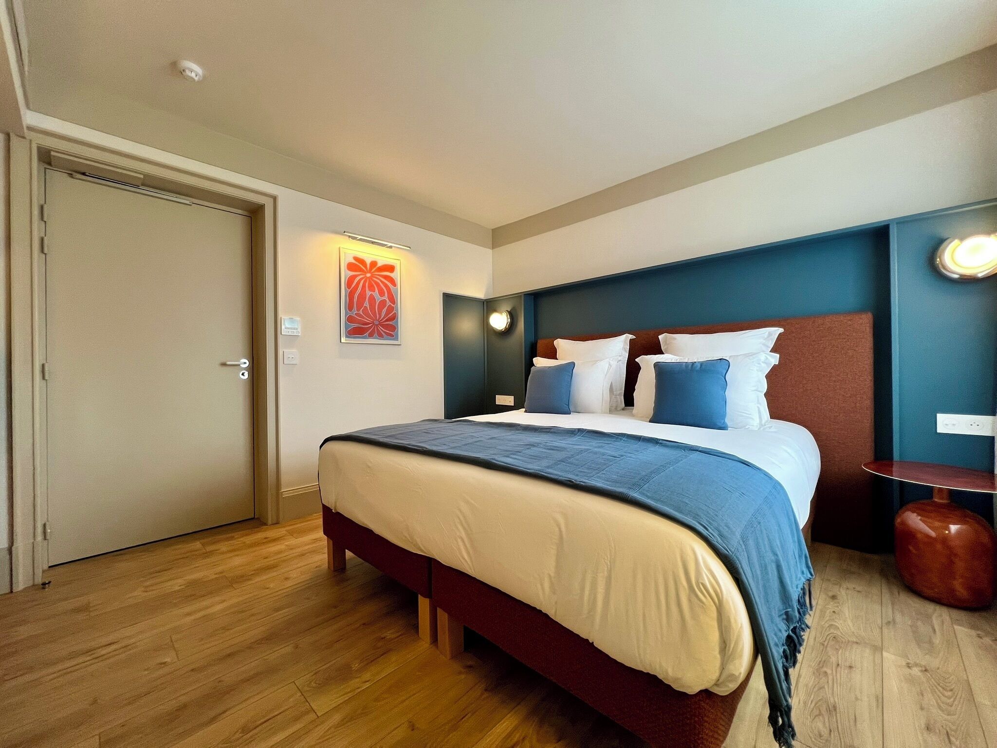 A hotel room with a bed, a door, a wall-mounted light, and a small table.