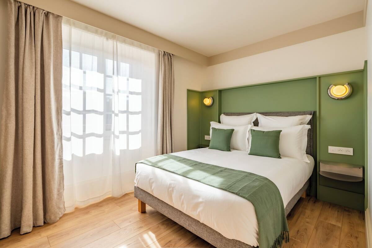 A hotel room with a bed, curtains, a nightstand, and a wall-mounted light.