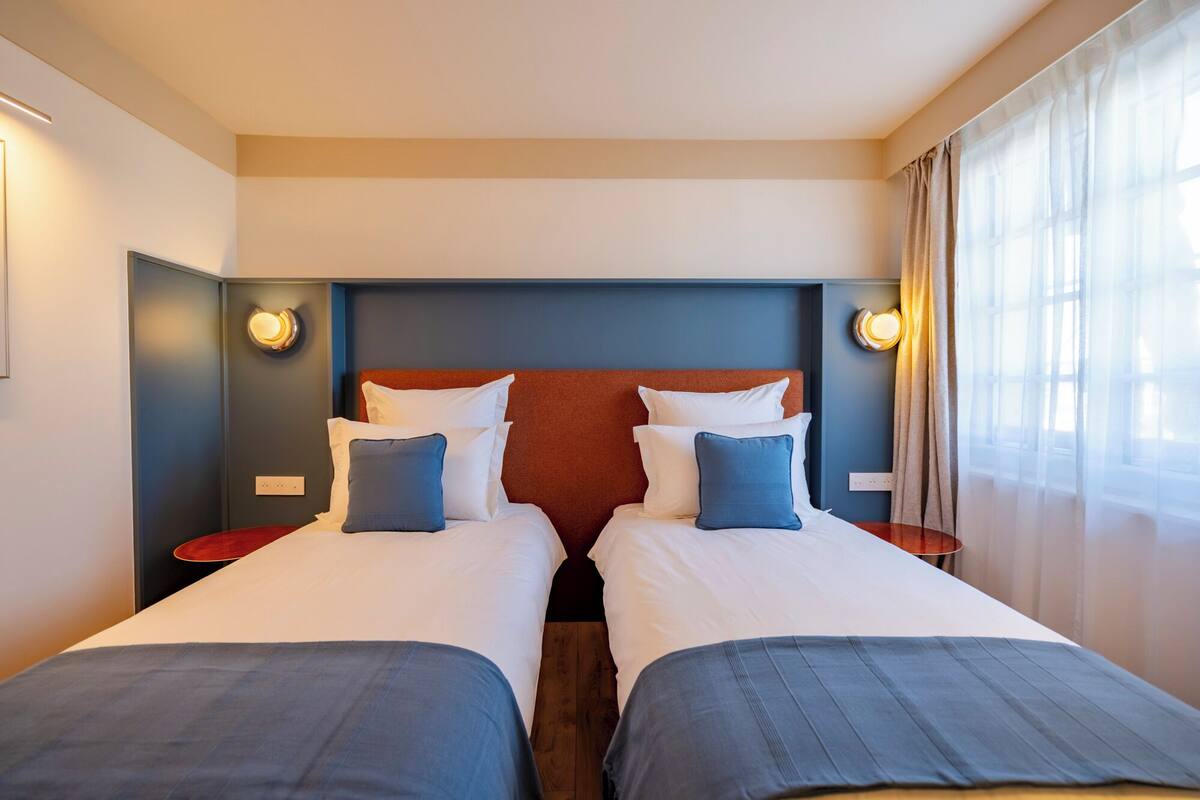 A hotel room with two beds, a wooden headboard, bedside lamps, and a window with curtains.