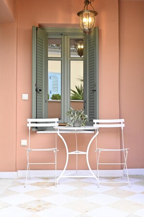 Une table en métal blanc, accompagnée de deux chaises, d’une plante en pot et d’un luminaire suspendu, devant une porte aux volets verts.