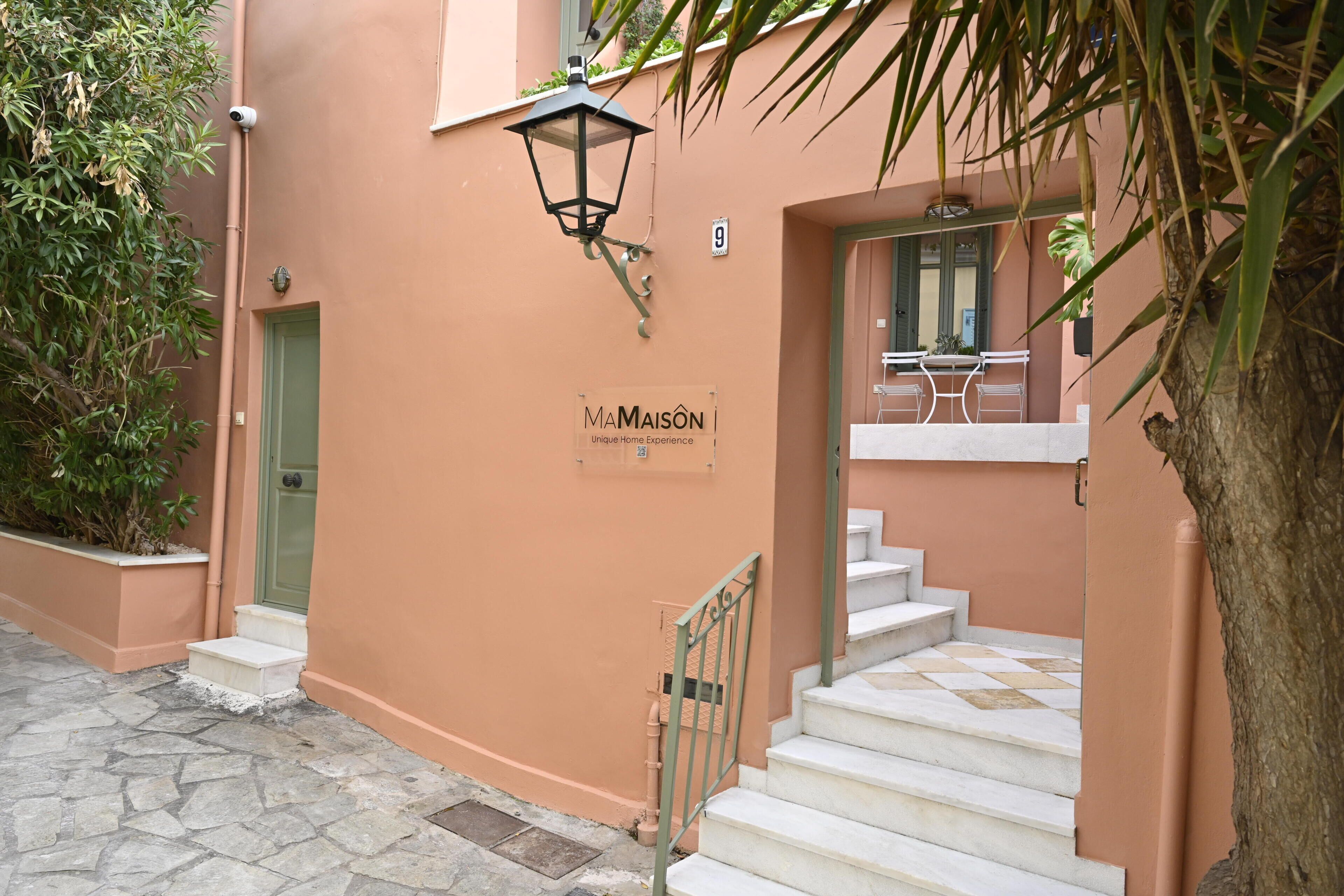 A building with a sign reading 'MAMAIOSON Unique Home Experience,' a staircase, and a security camera.