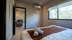 Une chambre dotée d’un grand lit, d’une fenêtre avec stores, d’un appareil de climatisation et donnant sur un espace de vie.