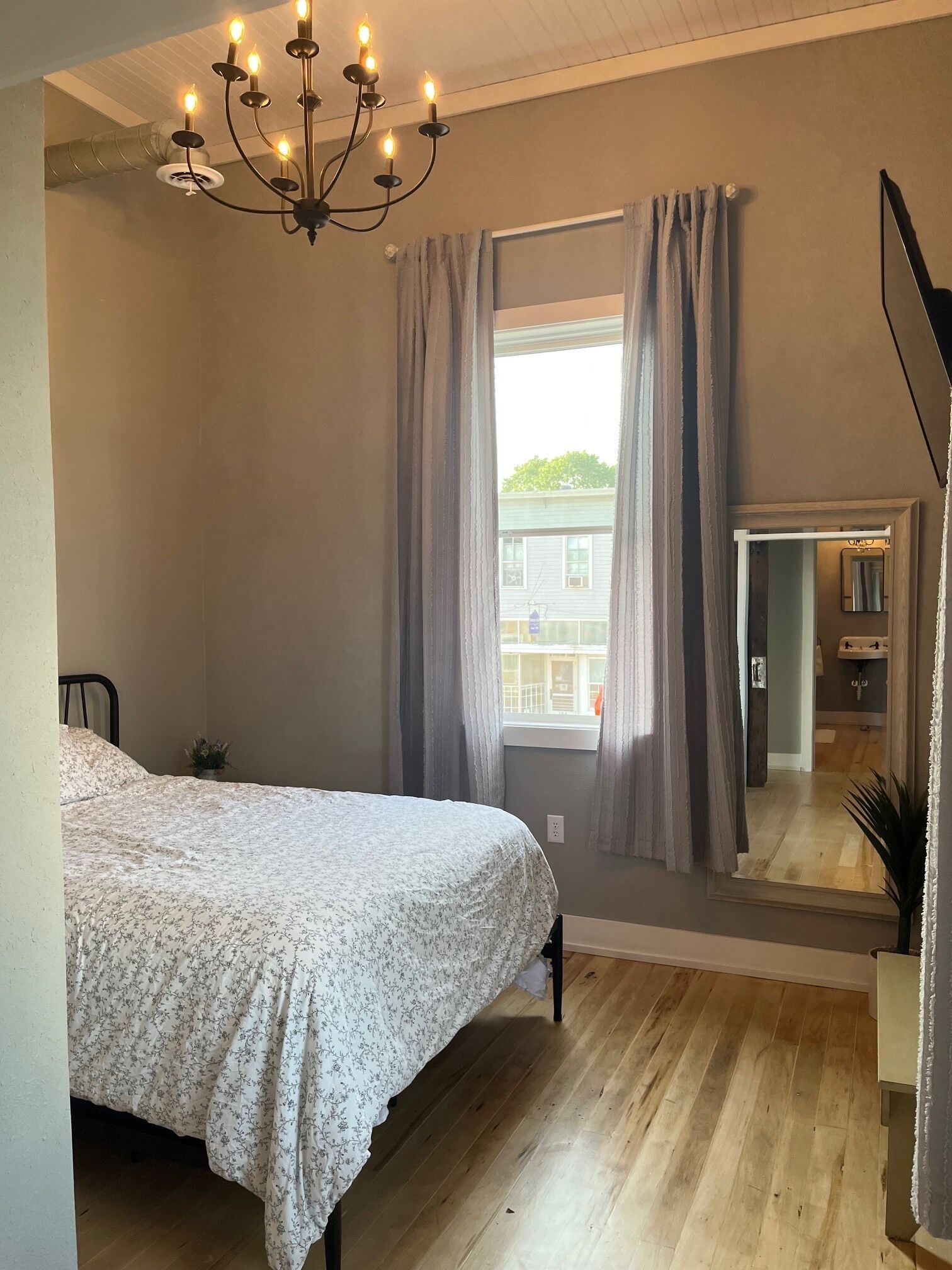 A bedroom with a bed, a chandelier, a window with curtains, and a mirror reflecting the bathroom.