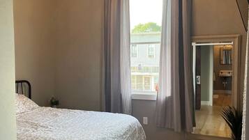 A bedroom with a bed, a chandelier, a window with curtains, and a mirror reflecting the bathroom.