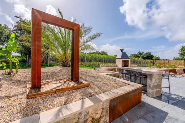 An outdoor patio with a wooden frame structure, seating area, and a small building in the background.