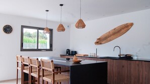 Una cocina moderna con una isla central, lámparas colgantes de mimbre y un detalle decorativo de tabla de surf de madera, además de un comedor con sillas de mimbre.