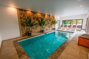 An indoor swimming pool with a stone wall, potted plants, and lounge chairs.