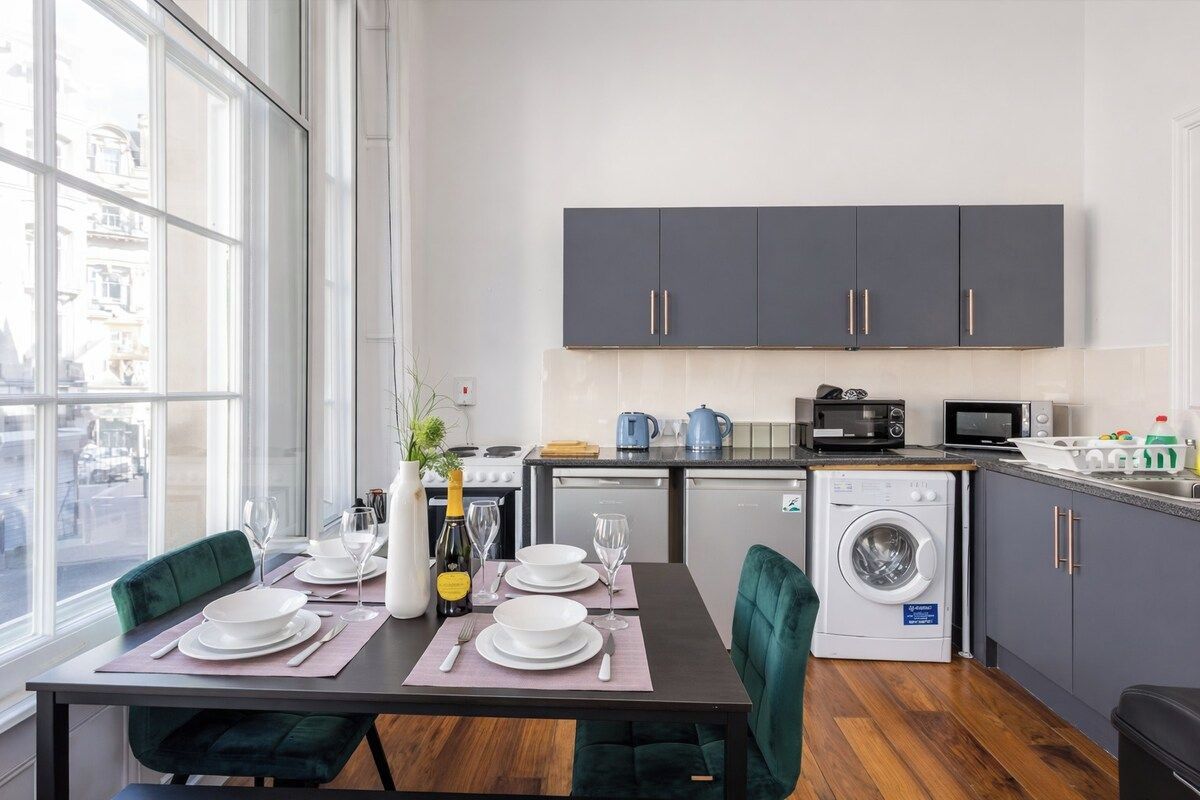 Une cuisine moderne avec des meubles gris, un lave-linge et un coin repas avec une table mise pour deux personnes.