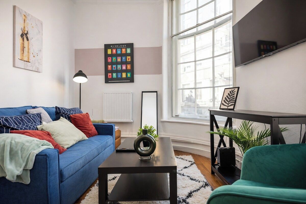 A modern living room with a blue sofa, a black coffee table, and a green armchair. There is a large window providing natural light and a view of the outside.