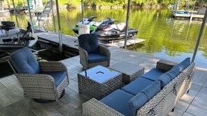 Móveis de vime externos com almofadas, uma mesa de vidro e vista para um cais com barcos.