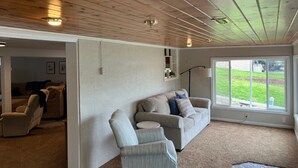 Uma sala de estar com teto de madeira, um sofá bege, uma poltrona cinza e uma janela grande com vista para um gramado.