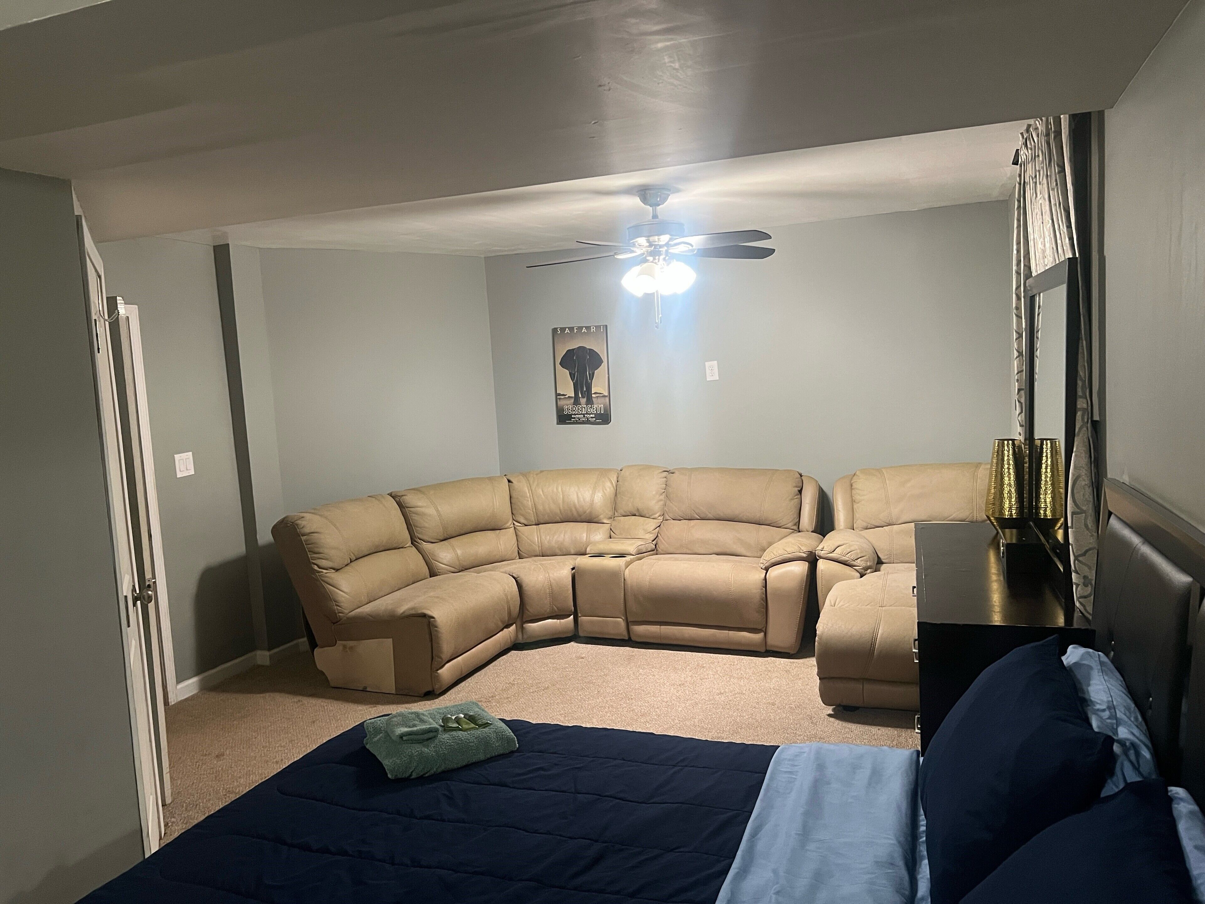 Una habitación con un sofá seccional, una cama con colcha azul y blanca, un ventilador de techo y un cartel en la pared.
